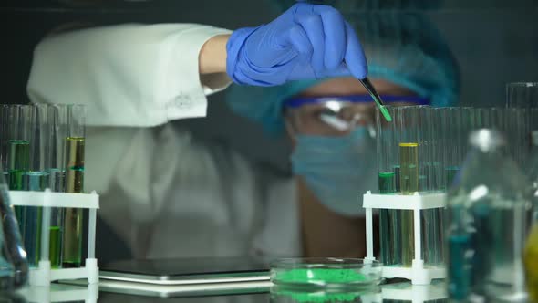 Lab Assistant Adding Green Powder to Tube and Typing Result on Tab, Technology