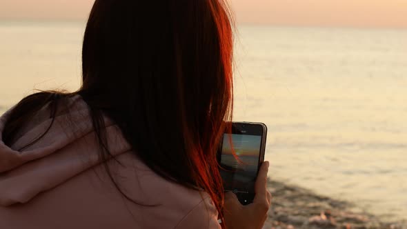 A beautiful girl photographs the dawn of the sun on the phone. The girl rejoices at the sunrise.