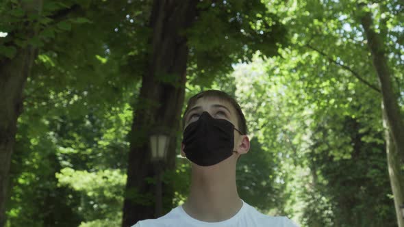 Young man in medical protective black mask in a forest with green foliage and trees 
