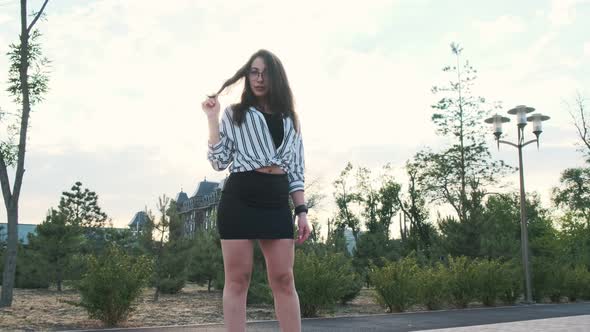 Beautiful girl in black high-heeled shoes, a black skirt and a striped shirt is standing in the park