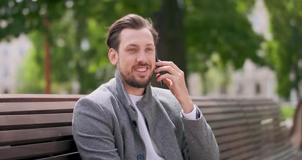 Young Man with Mustaches and a Beard is Sitting on a Bench in the Square Speaking By the Smartphone