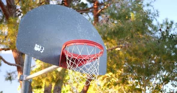 An old basketball hoop with a metal backboard, orange rim and net in an empty park court at sunrise.