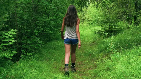 A tall brunette woman walking down a green, grassy forest path lined with tall trees into the distan