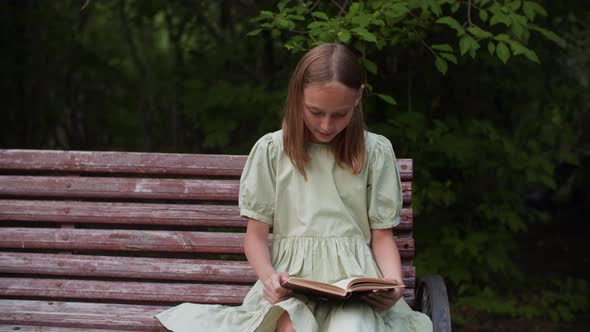 Teenager Girl Reading Book on Bench on Green Trees Background in Park