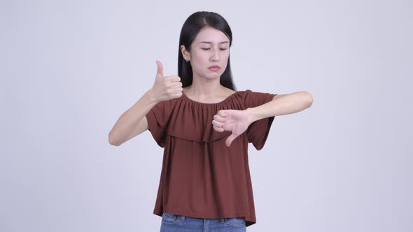 Confused Asian Woman Choosing Between Thumbs Up and Thumbs Down
