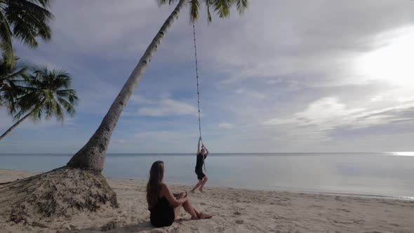 Young caucasian man rope swinging from palm tree on beach with young woman sitting in the sand watch