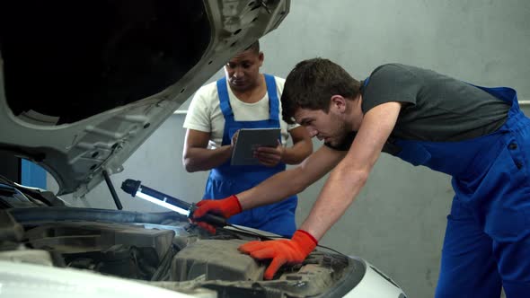 Mechanic with Flashlight Examines the Motor of Car and His Collegue Takes Notes