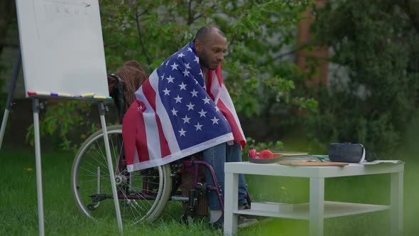 Sad African American Man Covered with USA Flag Sitting in Wheelchair on Summer Backyard Outdoors