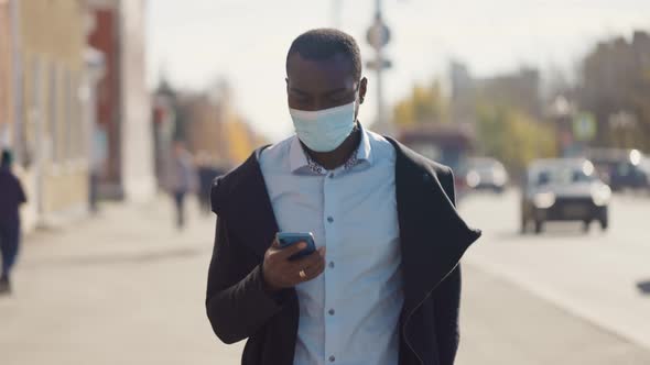 Portrait of African American Business Man in Mask, Smartphone in Hands. City Walk Down the Street