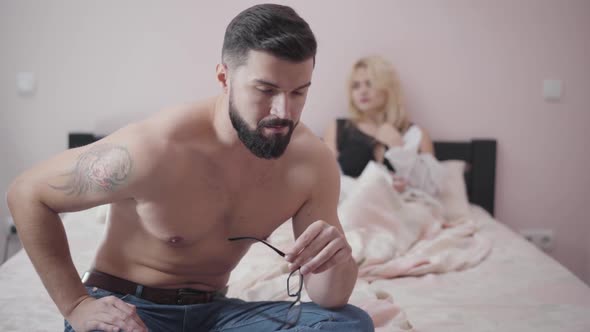 Close-up Portrait of Young Bearded Caucasian Man with Tattoo on Shoulder Sitting on Bed, Taking Off