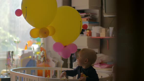 A baby girl standing in a crib and playing with balloons