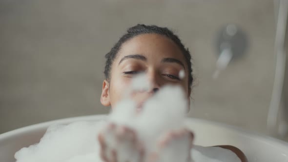 Young Happy African American Woman Relaxing in Foamy Hot Bath Blowing Foam at Camera and Smiling