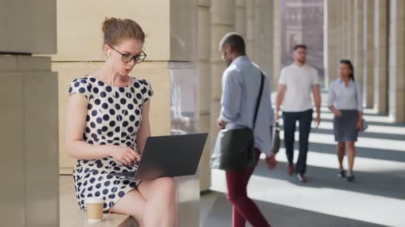 Young Businesswoman in Earphones Drinking Coffee Having Video Call on Laptop Sitting at City