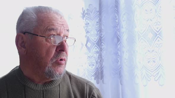 An elderly pensive man looks out the window of his house.