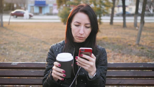 Beautiful Woman Use Phone Sitting on a Park Bench with Coffee Cup.