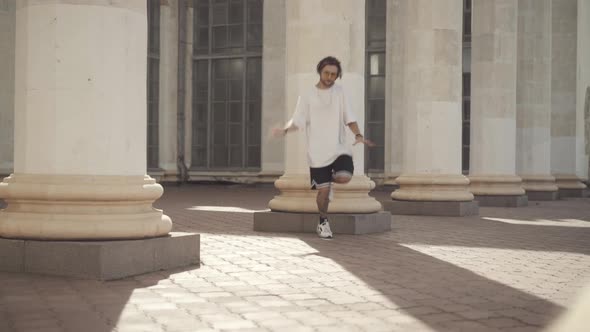 Wide Shot of Tattooed Man with Dreadlocks Dancing in Sunlight with Urban Building at Background