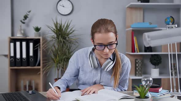 Blond Attractive Girl in Glasses Sitting at the Table and Writting Essay Using Book