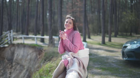 Portrait of Smiling Brunette Caucasian Woman with Tea Sitting on Tree Trunk in Sunshine