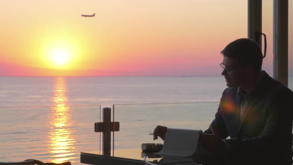 Smoking Businessman with Pad in Seaside Cafe at Sunset