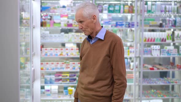 Unsure Caucasian Senior Man Choosing Pills in Pharmacy Leaving with Medication Bottle Rubbing