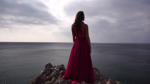 Side View a Young Beautiful Sensual Woman in a Red Long Dress Posing High Above the Sea on a
