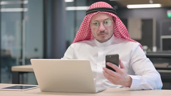 Middle Aged Arab Man with Laptop using Smartphone in Office