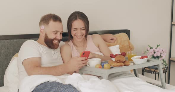 Happy Young Couple Spending Time and Laughing in Bed. Man and Woman Looking at Smartphone Screen and