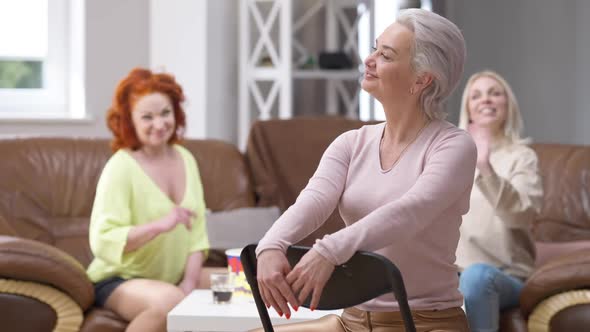 Confident Gorgeous Adult Woman Sitting on Chair at Home Looking Back at Friends Waving and Mocking