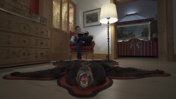 A man reads a book in a chair with a bearskin rug at a ski resort.