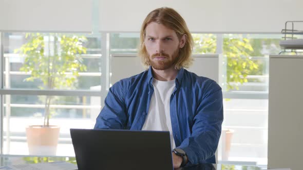 Happy Caucasian Young Businessman in Casual Clothes Sitting in Front of Laptop and Looking at Camera