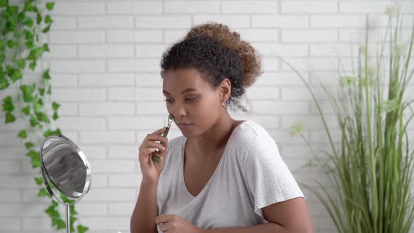 Young woman massaging face with jade roller while looking in mirror