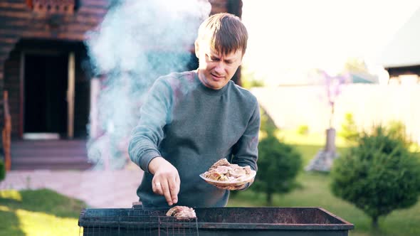 Young Man Stands on the Background of a Summer House Lays Marinated Pork on a Barbecue Grill