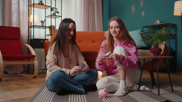 Girls Friends Siblings Sitting on Floor and Take Turns Dropping Dollar Banknote Into Piggy Bank