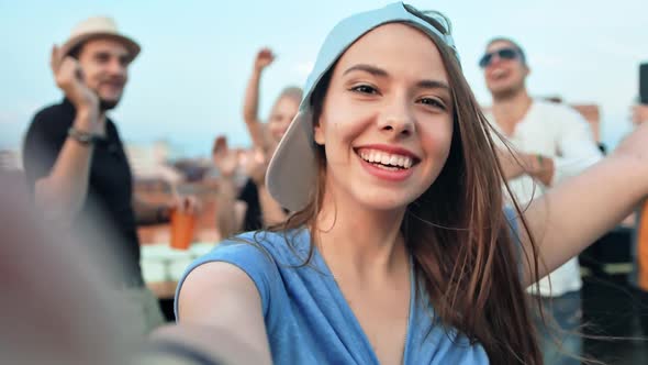 Closeup Face of Young Woman in Cap Taking Selfie Surrounded By Friends at Rooftop Party