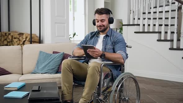 Guy in Headphones Sitting in Wheelchair in Living-Room and Listening Music from i-pad
