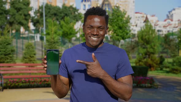 Smiling AfroAmerican Man Finger Points Smartphone Display with Green Screen