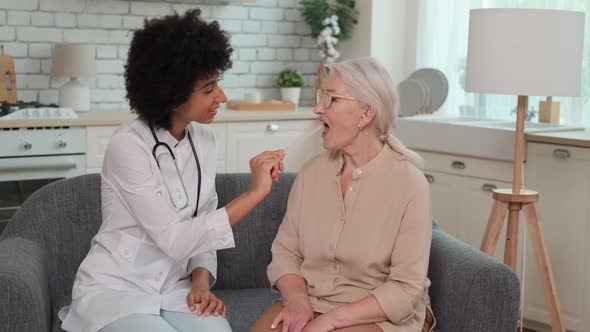 Afro American Woman Doctor Looking Throat Senor Woman Sitting on Sofa at Home