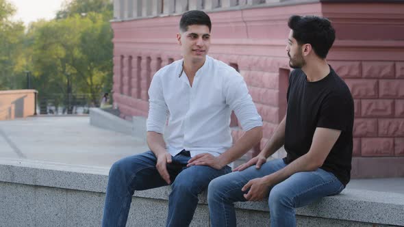 Two Positive Indian Arab Men Sitting Outdoors Having Conversation Discussing Educational Issues or