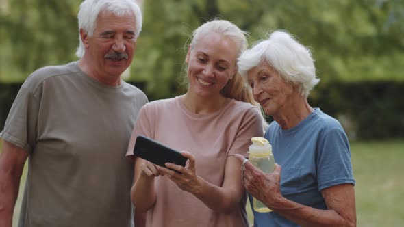 Cheerful Female Coach Using Smartphone and Chatting with Senior Couple Outdoor