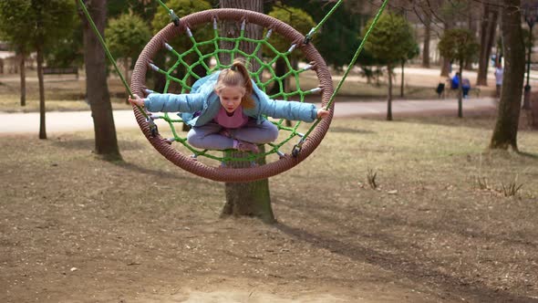 A Little Girl in a Blue Coat Rides on a Swing on Playground in Park