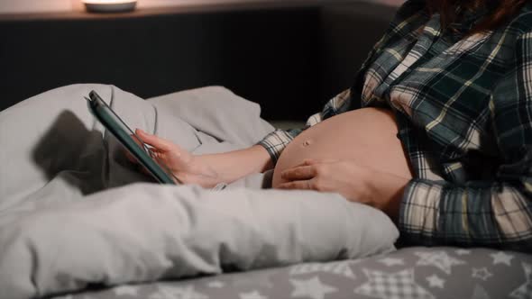 Pregnant with tablet computer in bedroom showing and touching belly.