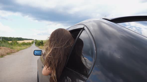 Beautiful Woman Leaning Out of Car Window and Enjoying Trip While Riding Through Country Road. Young
