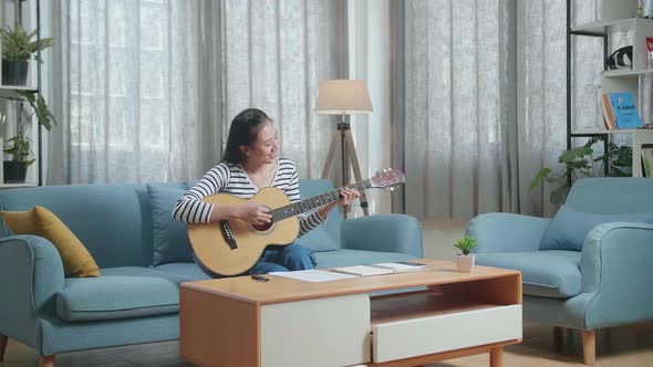 Smiling Asian Woman Composer With Paper And Notebook On Table Playing Guitar At Home