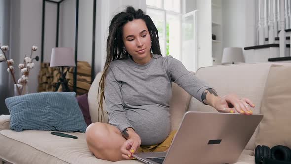 Pregnant Woman with Dreadlocks Sitting on the Soft Couch at Home and Starting to Work on Laptop