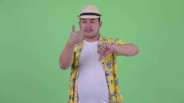 Confused Young Overweight Asian Tourist Man Choosing Between Thumbs Up and Thumbs Down