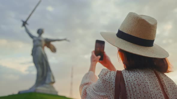 An attractive woman in a hat near Mother Motherland taking a photo on a smartphone on Mamayev Kurgan