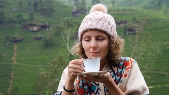 Young Woman In Knit Hat Smelling Tea In Cup Outdoors At Tea Plantation Hills