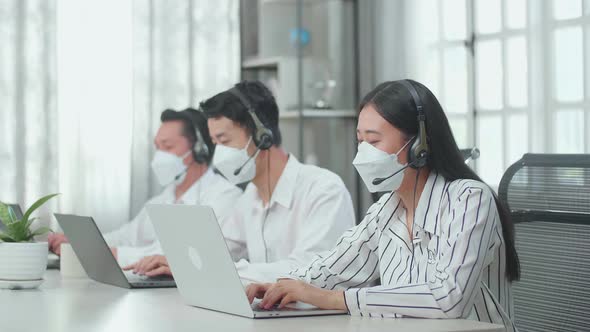 Three Asian Call Centre Agents Wearing Masks Typing On Computers While Speaking To Customers