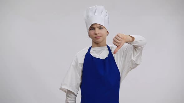 Caucasian Boy in Blue Apron and White Chef Hat Posing at Background Showing Thumb Down