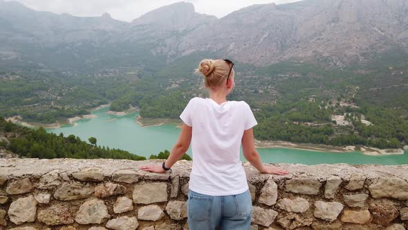 A Young Girl Looks at a Beautiful View with a Blue Lake and Mountains From the Wall of an Old Castle